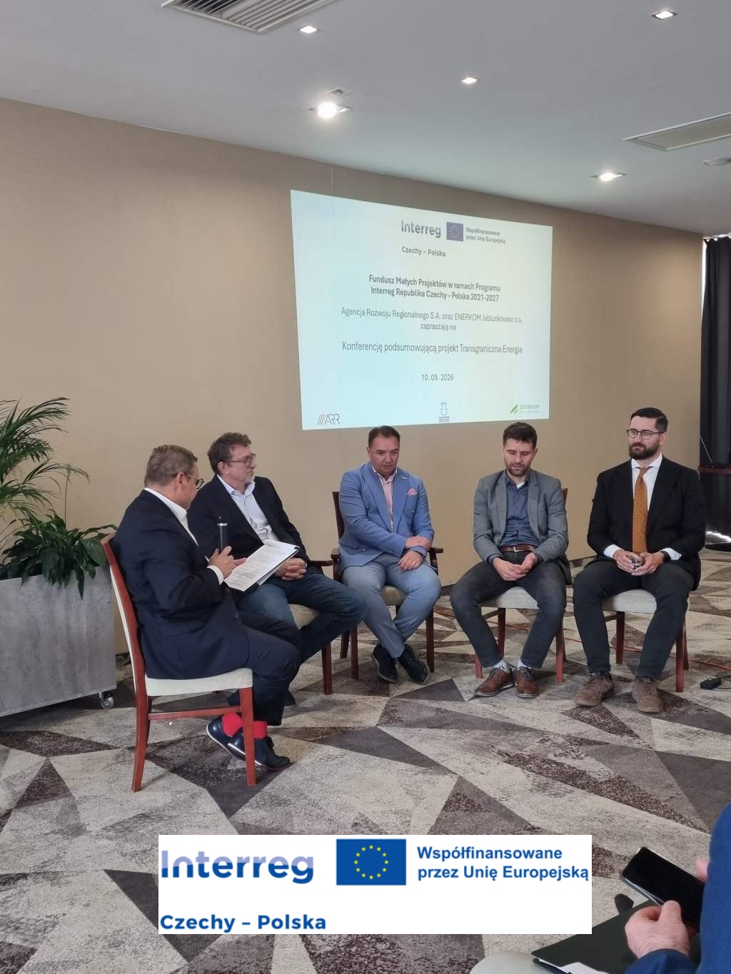 Panel dyskusyjny podczas konferencji podsumowującej projekt „Transgraniczna Energia”. Pięć osób siedzi na krzesłach w sali konferencyjnej, a w tle wyświetlony jest slajd z informacją o wydarzeniu realizowanym w ramach programu Interreg Czechy–Polska 2021–2027. Na zdjęciu widoczne jest również logo Interreg oraz informacja o współfinansowaniu przez Unię Europejską.